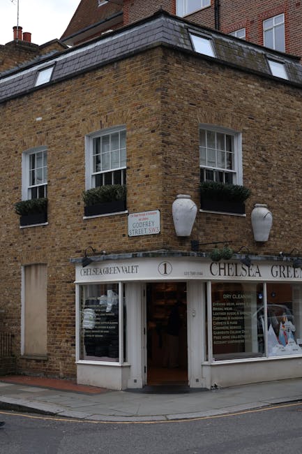 A three-story corner building constructed of brown brick with a flat roof features two white-framed sash windows on the upper floor, each fitted with black flower boxes containing green plants. Attach