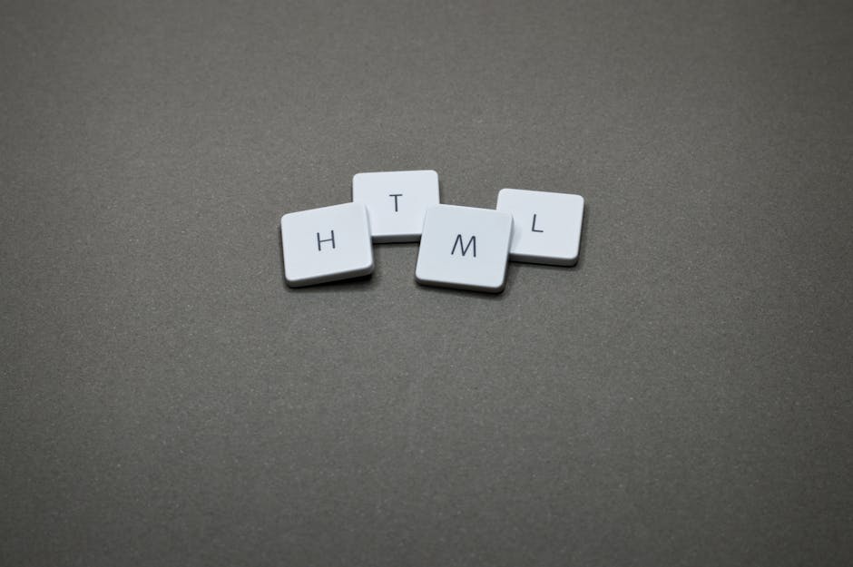 Four white scrabble tiles with black uppercase letters spell out the word 'HTML' and are arranged on a smooth, matte gray surface. The tiles are positioned in a slightly irregular formation, with the 