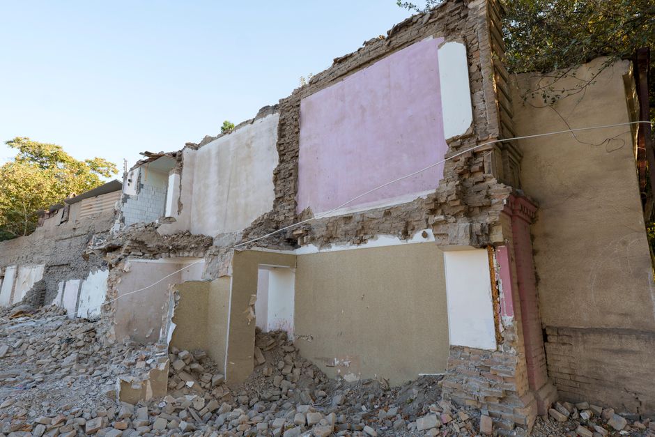 The image depicts a partially demolished brick building exterior, with sections of the wall torn away to reveal interior painted walls in white and light pink, surrounded by a large pile of rubble, br