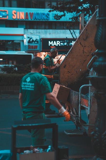 A man wearing a face mask, blue shirt, and gloves is standing on the edge of a black waste collection truck, loading and sorting various household furniture and large fixtures, including a wooden cabi