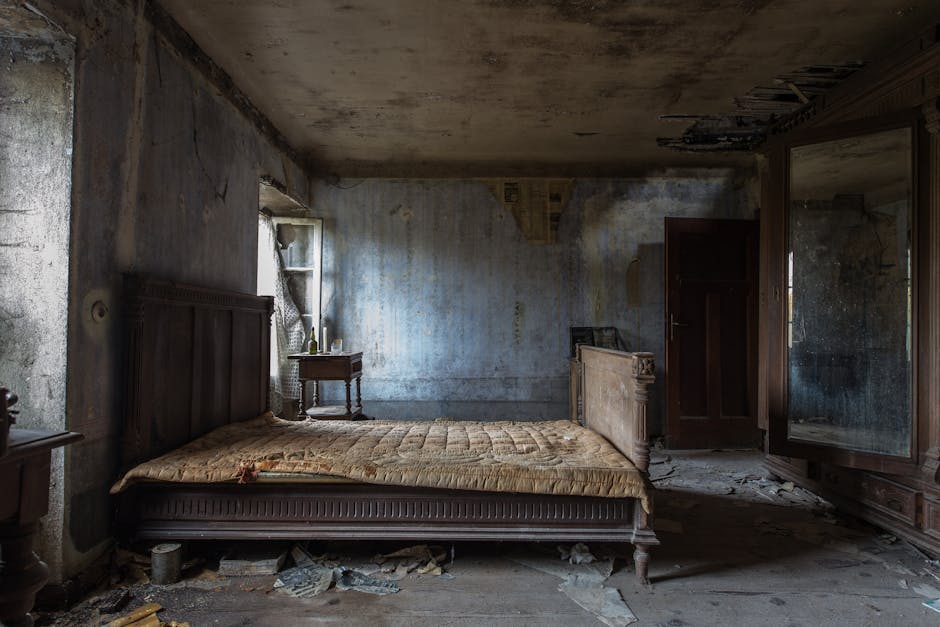 An empty bedroom with white walls, featuring a double bed with a wooden headboard and a floral mattress, flanked by two small matching bedside tables with a wood finish and black metal legs. To the ri