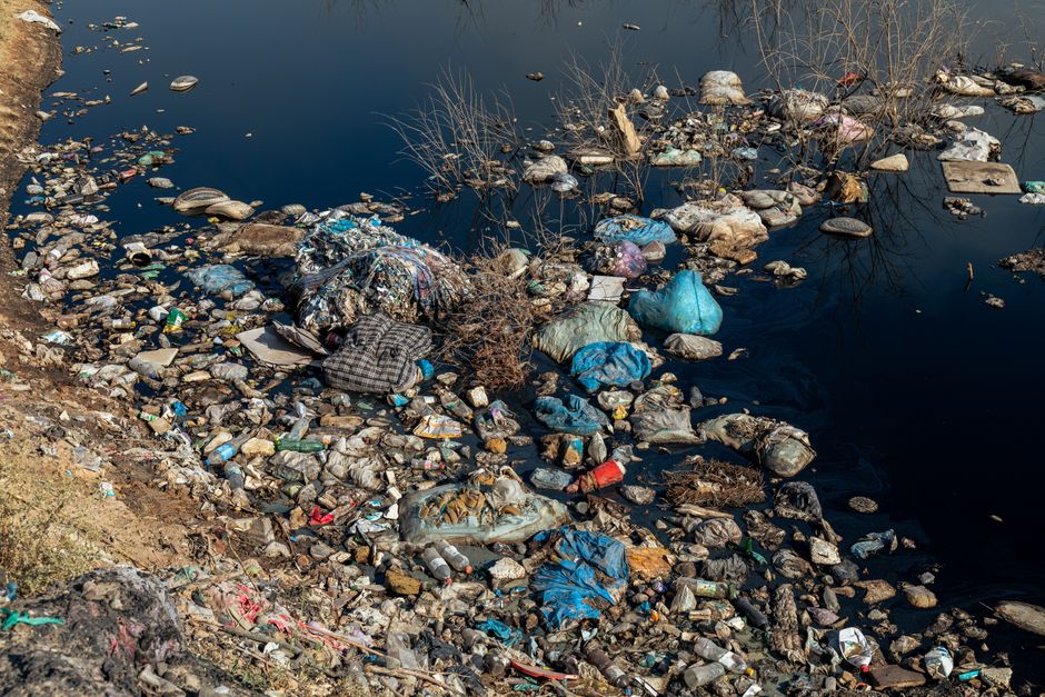 An expansive view of a large landfill site filled with a dense layer of mixed waste materials, primarily consisting of crumpled paper, plastic debris, food packaging, and discarded household items. Th